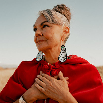 Model looks to the left with hands at their chest wearing the mussel-inspired earring "Coastal Strata" in silver mirror acrylic by Copper Canoe Woman.