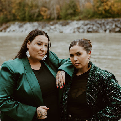 Two models stand together facing the camera while wearing "Echoed U" earrings from Copper Canoe Woman, the left in white acrylic and right in black acrylic.