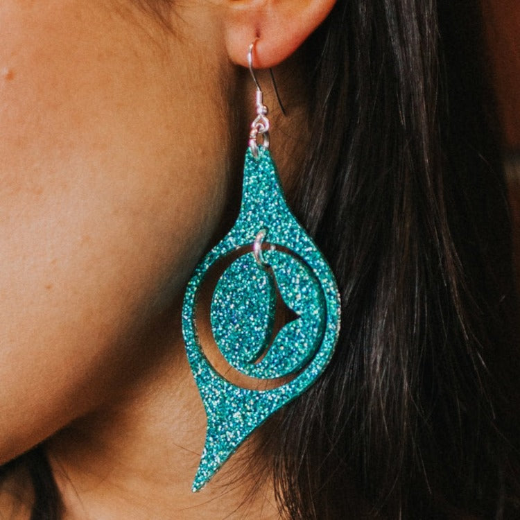 A closeup of an "Octopus Eye" earring in holographic teal glitter acrylic from Copper Canoe Woman.
