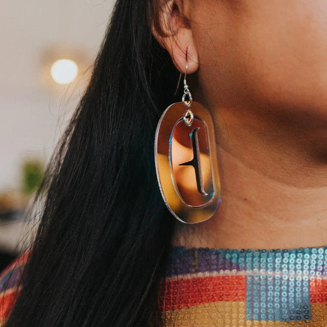 A close-up image of a person's ear wearing a large ovoid earring in iridescent acrylic with a cutout design. 