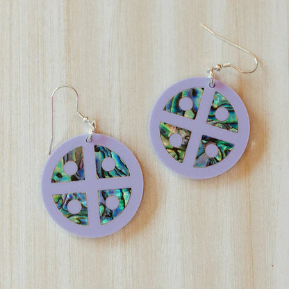 Close up of Copper Canoe Woman "Protector Shield" earrings in lavender acrylic with abalone inlay sitting on a table.