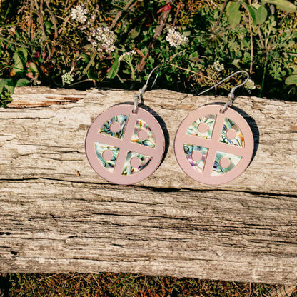 A pair of rose gold mirror acrylic "Protector Shield" earrings from Copper Canoe Woman sit on a log.
