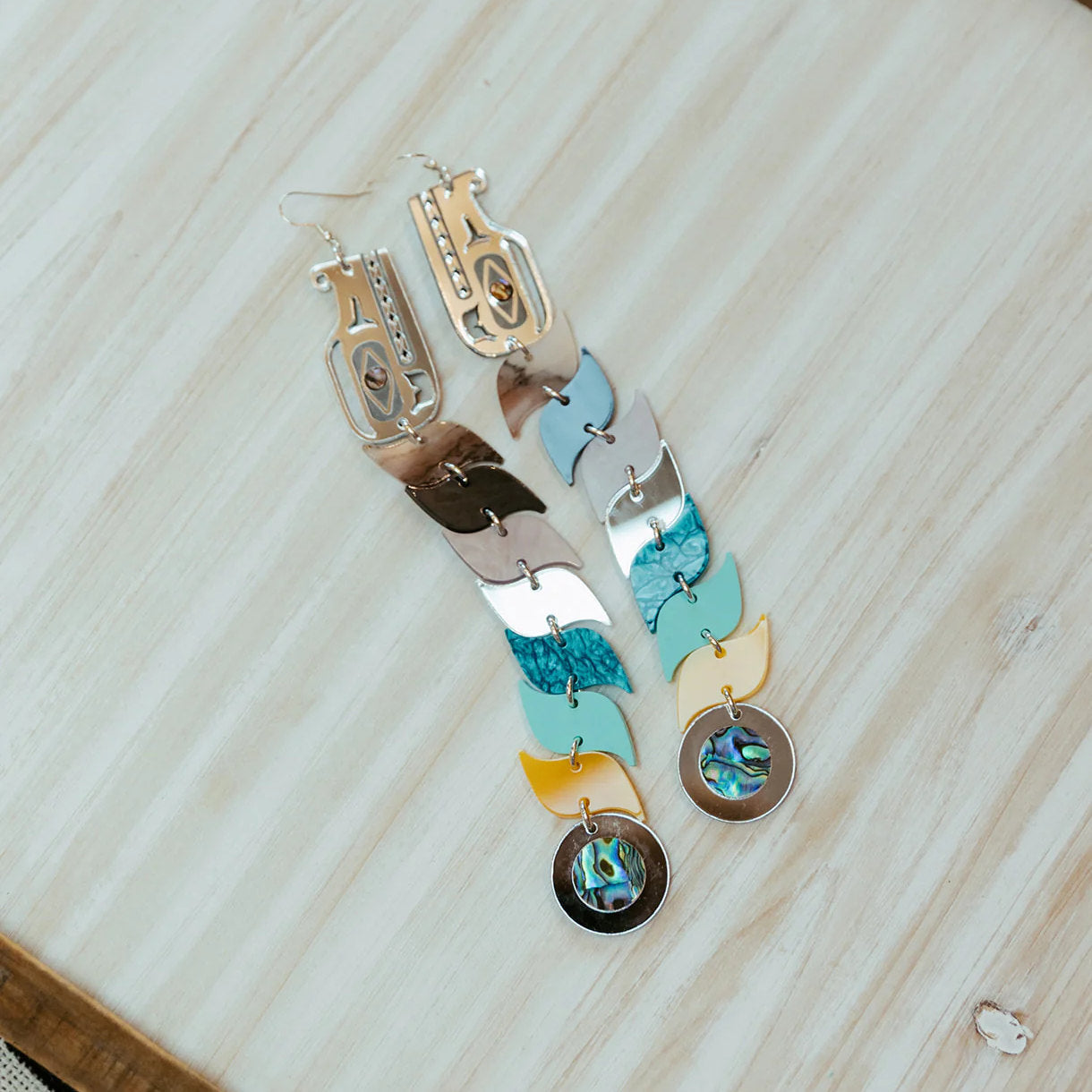 A pair of blue and silver mirror acrylic serpent earrings resting on a wooden table from Copper Canoe Woman.