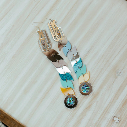 A pair of blue and silver mirror acrylic serpent earrings resting on a wooden table from Copper Canoe Woman.