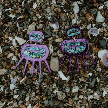 A rain covered pair of purple mirror acrylic Urchin earrings from Copper Canoe Woman laying on pebbles.