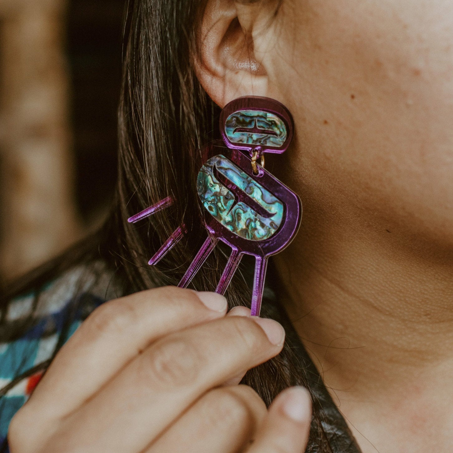 Closeup of model wearing and holding a pair of purple mirror Urchin earrings from Copper Canoe Woman.