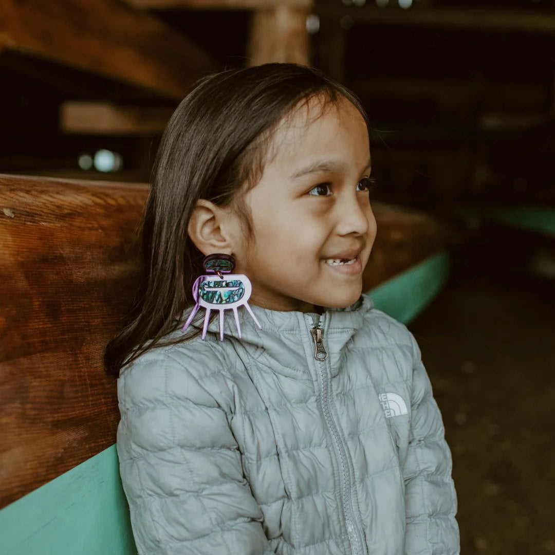 A youth grins to something off camera while wearing purple mirror acrylic Urchin earrings from Copper Canoe Woman.